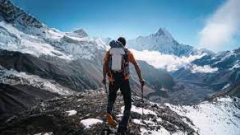 Still from Race to the Summit: White rock climber walking on a mountain top with the use of ski trail poles with back to the camera, wearing an orange-colored T-shirt, dark pants, dark hiking boots and a white, orange, and black backpack