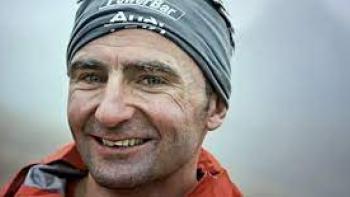 Still from Race to the Summit: Record-breaking White Swiss mountain climber Ueli Steck smiles at the camera, wearing an orange parka and gray sports bandana headband with words in white letters