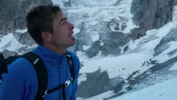 Still from Race to the Summit: Right facial profile of record-breaking White Swiss rock climber Dani Arnold standing on a snow-covered mountain looking upward, wearing an electric blue hardshell jacket and carrying a black backpack