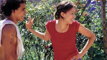 Raising Victor Vargas movie: Two young people laughing together outdoors near a chain-link fence with greenery in the background.