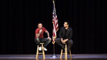 Refuge Movie: Chris Buckley (left) and Heval Kelli are on a stage, sitting on stools, an American flag between them