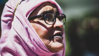 Refuge Movie: Mama Amina, wrapped in a pink head scarf and wearing glasses, looks to viewers' right