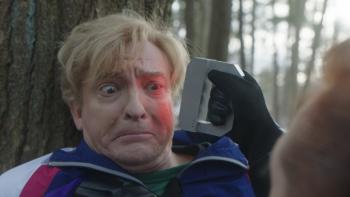Relax, I'm from the Future: Rhys Darby against a tree with a gray object held to his head that’s shining a red light