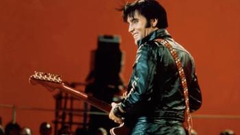 Return of the King: Elvis Presley stands smiling over his shoulder against a dark orange backdrop holding a guitar and wearing a black leather suit.
