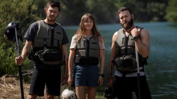 River Wild movie: In front of river behind them, a white woman center is with white man on right and white man on left, with everyone wearing black life jackets