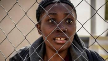 Run: A Black woman looking through a chain-link fence, appearing worried
