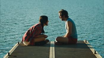 Sacramento movie: Maya Erskine and Michael Angarano laugh while facing each other sitting on a dock by a lake