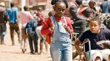 Safari: Black Kenyan girl center walks with Black Kenyan wheelchair user boy on right, down a Kenyan street with Kenyans in the background