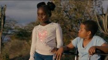Safari: Black Kenyan girl in white shirt center walks in outdoors alongside Black Kenyan wheelchair user boy in light blue t-shirt 
