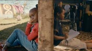 Safari: In split view, a Black Kenyan girl in jeans and red shirt sits outside back against wall, while on other side of wall sits a Black Kenyan boy in dark blue shirt and tan shorts with paper in his hands