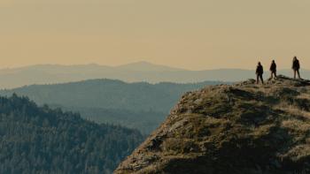 Sasquatch Sunset Movie: Three Sasquatch stand together on the top of a hill, overlooking a tree-filled valley