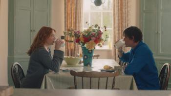 She Is Love: A White woman and man sit at a breakfast table. Both are drinking a glass of milk