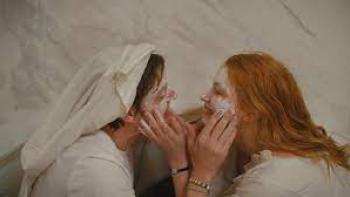 She Is Love: A White man and woman rub white cream into each other's faces, dressed in white, against a white background