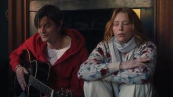 She Is Love: A White man sits against the living room wall playing guitar, with a White woman sitting next to him