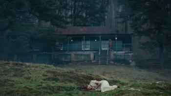 She Will: A figure in while lies on the grass in front of a cabin.
