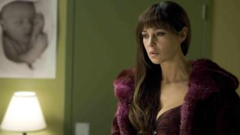 Monica Bellucci wears a purple dress with matching fur coat and stands in a dimly lit bedroom.