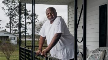 Silver Dollar Road movie: Black man in white t-shirt center stands leaning over rail on front porch of white house with grass and shed in distance