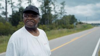 Silver Dollar Road movie: Black man in white t-shirt and black baseball cap left stands next to two lane road with grass and trees on left side