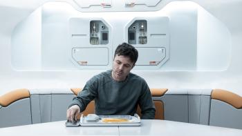 Slingshot Movie: Seated at a gray table, John (Casey Affleck) eats a lonely meal