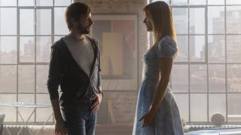 Soulm8te movie: Standing in front of picture windows, David, in a hoodie, speaks to the robot Sara, who's wearing a blue dress