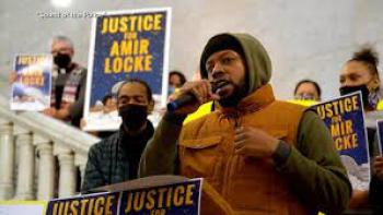 Still from Sound of the Police: Ron Adams, a Black community organizer holding a microphone and addressing an audience at Minneapolis City Hall after the death of Amir Locke by police in 2022, surrounded by BIPOC and White activists carrying signs that read "Justice for Amir Locke,"  and wearing black knit cap, dark green hoodie, and brownish-orange vest jacket