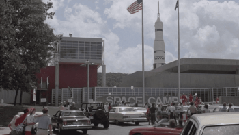 SpaceCamp movie: Space Camp center entrance with cars, flags, and a rocket in the background.