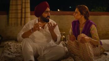 Saunkan Saunkne: While sitting on a mattress, an Indian man in a turban talks to an Indian woman