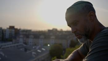 Stand: Black man right looks out over rooftops as sun sets
