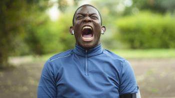 Street Flow 2 movie: Black French bald man in blue jumper wearing earbuds opens his mouth and yells while outside in a park with trees and bushes behind him