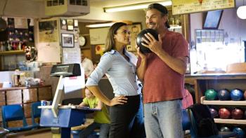 Paula Patton and Kevin Costner stand in a bowling alley. Costner holds a bowling ball to his chin and is smiling.