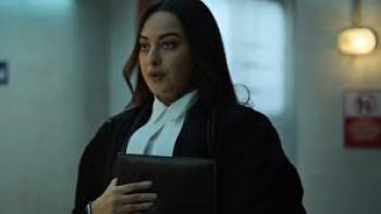 System movie: Indian woman in formal legal attire holding a folder, walking in a dimly lit hallway.