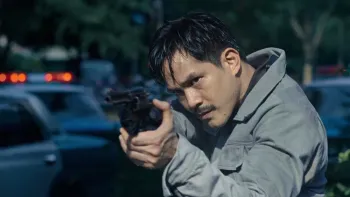 Tee Yai: Born to be Bad movie: Thai man in grey shirt center points pistol forward with police cars behind him