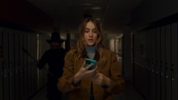 Thanksgiving Movie: Teen Jess (Nell Verlaque) stands in a dark hallway looking at her phone while a figure in a pilgrim hat, holding an axe, sneaks up behind her