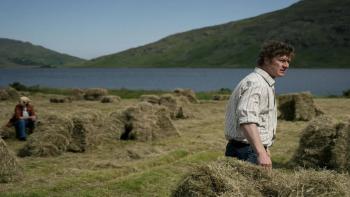 That They May Face the Rising Sun: A White man stands in a field while a White woman sits on a bale of hay