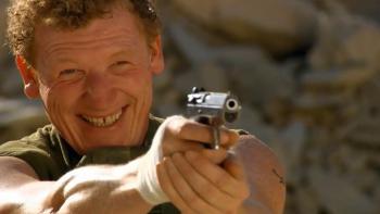 The Business: Man smiling while aiming a gun outdoors