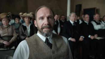 The Choral movie: A bearded Ralph Fiennes in vest and tie stands in crowded hall