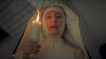 The Devils: Nun holding a lit candle, gazing forward, in a dimly-lit room