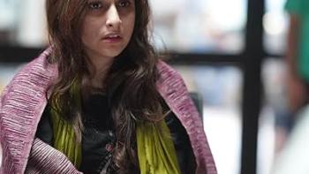The Diplomat: A woman with long dark hair wearing a pink cardigan and yellow top sits at a table with papers