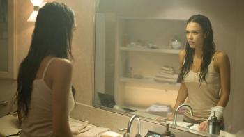 Jessica Alba stands in front of a mirror in a bathroom, wearing a tank top and a sober expression. The lighting is dusty and yellow.