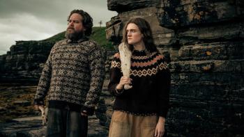 The Incomer: A man and woman in patterned sweaters stand by rocky cliffs under a cloudy sky
