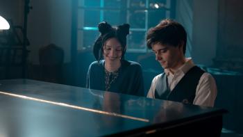 The Magic Flute Movie: A teen girl with black hair sits with and smiles at a teen boy, at a piano 