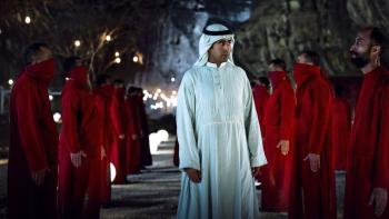 The Matchmaker: Saudi men in red robes line up on left and right while a Saudi man in a white robe and kaffiyeh walks down the center