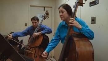 The Only Girl in the Orchestra: A woman and a man are shown from the waist up playing double basses together.