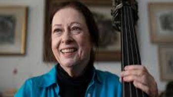The Only Girl in the Orchestra: A woman with brown hair pulled back wearing a blue shirt with her fingers on the neck of a double bass.