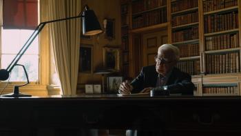 Still from The Pigeon Tunnel: John le Carré, the White British author who is the subject of the film, is seated at a desk with a black lamp while writing with a pen, the gray-haired writer is wearing a dark-colored sports jacket, a light-colored Oxford shirt with the collar opened, and gray pants, and in the background are bookshelves and framed images hanging on a wall or positioned on a nearby table
