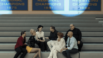 The Prom movie: Seven people sit talking on gym bleachers under championship banners.