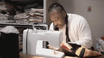 The Quilters: Chill, a man of color with long locks and grizzled beard, bends over a sewing machine preparing to seam some fabric.