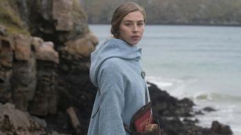The Road Dance: A young woman stands in a light blue coat on the coastline, the sea stretching out behind her