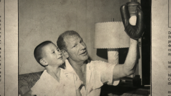 Still from The Saint of Second Chances: Mike Veeck (left), as a kid, looks upward along with his dad, baseball owner Bill Veeck, who holds a catcher's mitt with a white baseball in his raised left arm, both wearing white short-sleeved shirts unbuttoned at the top
