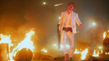 The Shadow Strays movie: Indonesian man in white suit center stands with large rifle and ammo belt in warehouse on fire